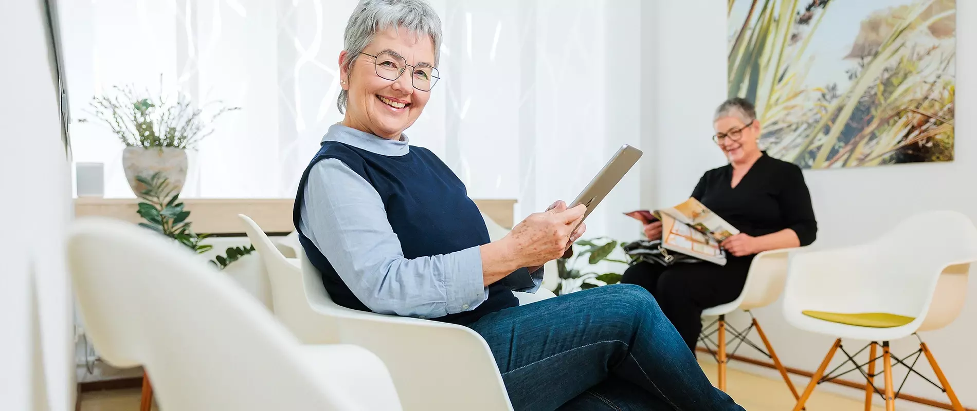Aufnahme einer im Wartebereich sitzenden Dame, die eine Zeitschrift in der Hand hält und in die Kamera lächelt.